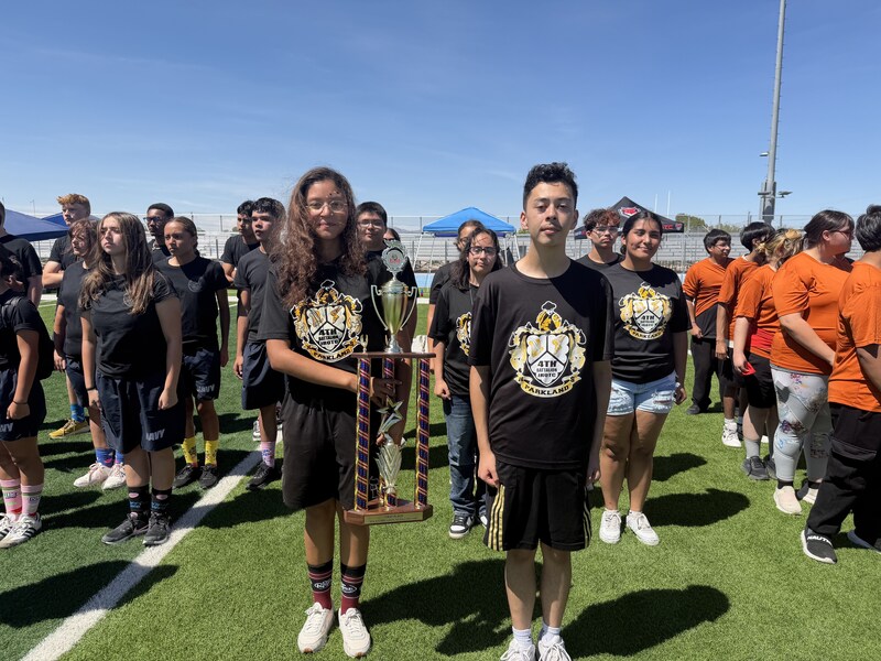 Two Parkland JROTC students stand at the front of a group on a sports field holding a large first-place trophy.
