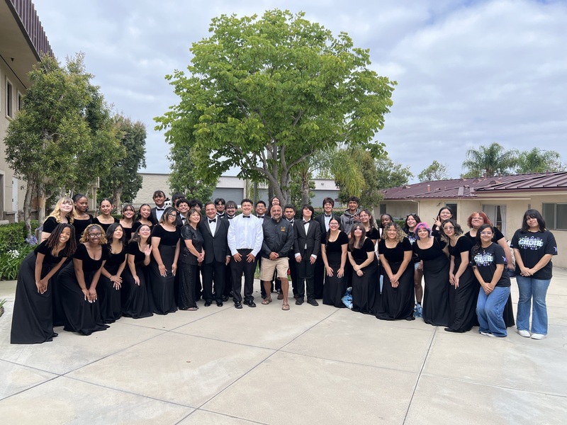 The choir members are dressed in formal black competition attire posing with the principal and sponsors outside. Buildings, trees and a cloudy sky are in the background.
