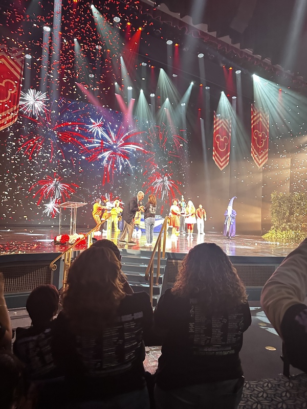 The choir teacher is accepting an award on a stage. The audience is standing and applauding.