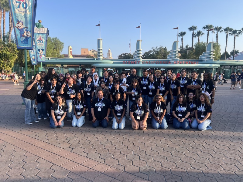 Choir students and chaperones pose for a picture wearing black t-shirts and jeans outside at an amusement park.