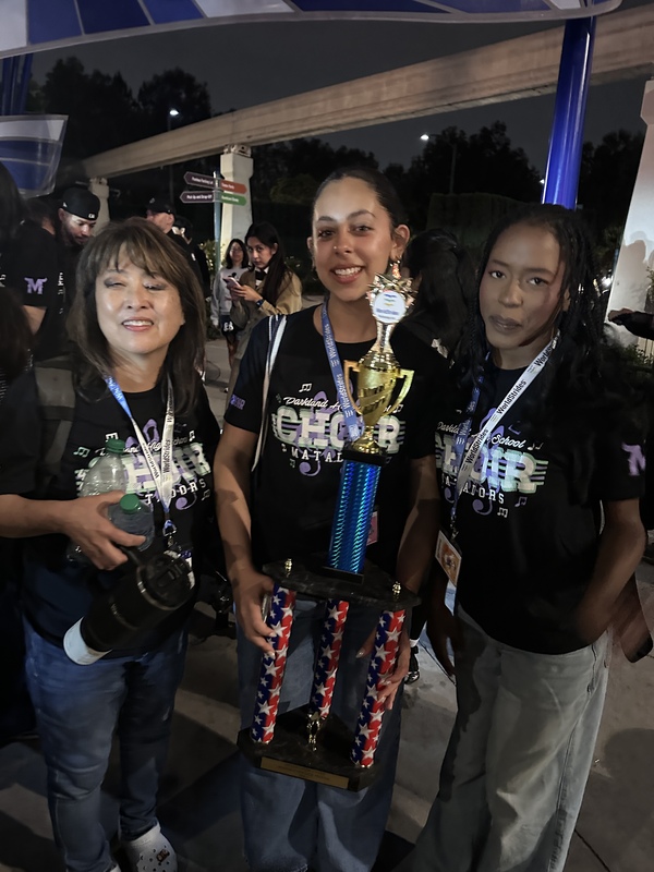 Choir teacher and 2 students wearing black t-shirts, holding a tall trophy.