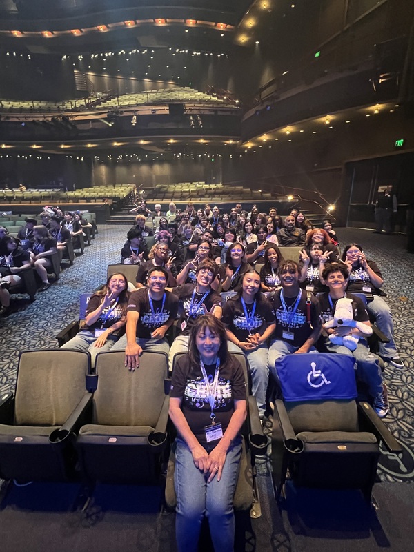 Choir and teacher wearing Choir school t-shirts sitting in theater seats.