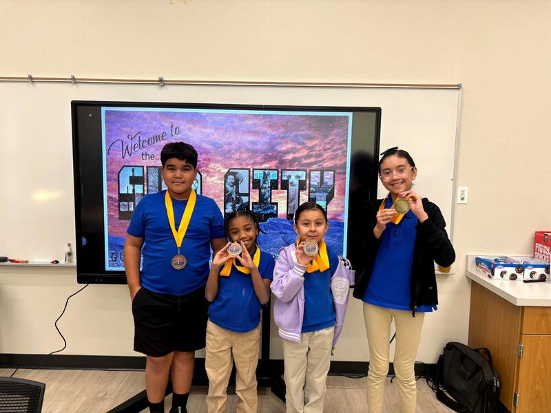 Four Q-BEC GT Challenge champions proudly display their medals in front of a colorful digital backdrop in a classroom.