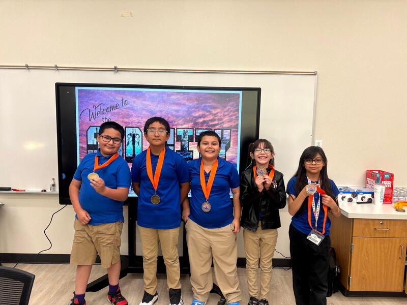 Four Q-BEC GT Challenge participants proudly display their medals in front of a colorful digital backdrop in a classroom.