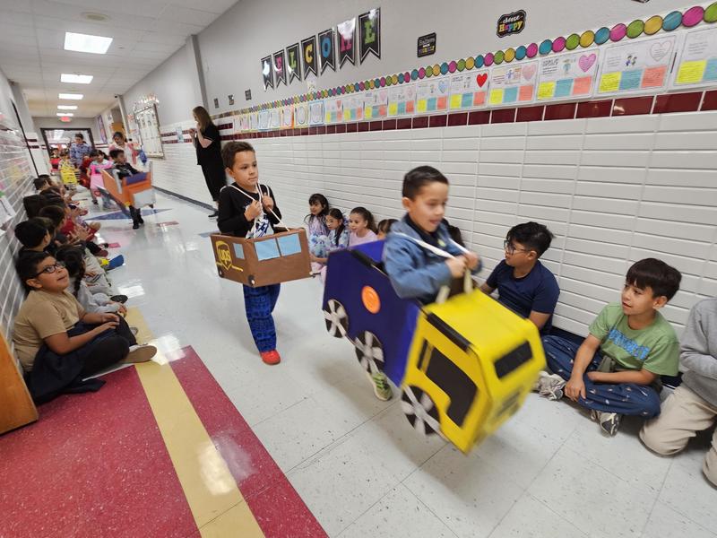 PK students walk with their garbage truck and a UPS  vehicle model around their shoulders