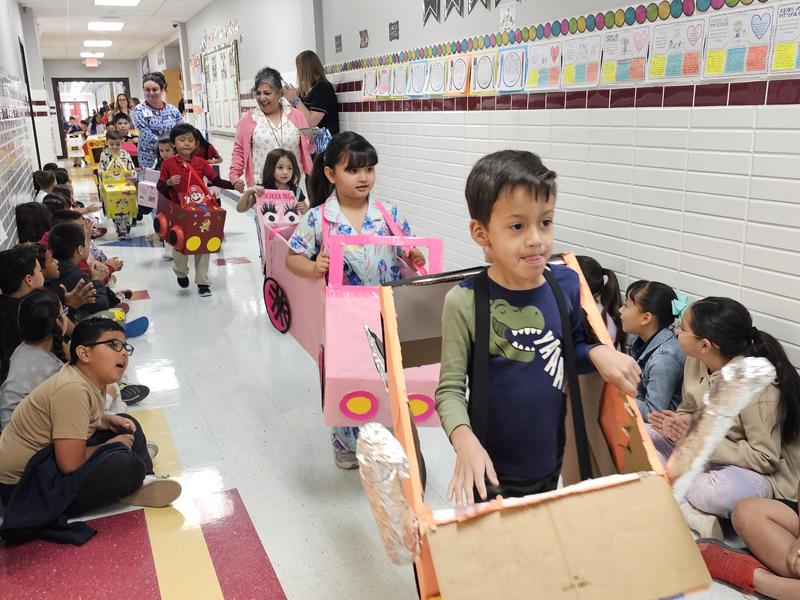PK students walk down the hallway in their colorful pink and red cars