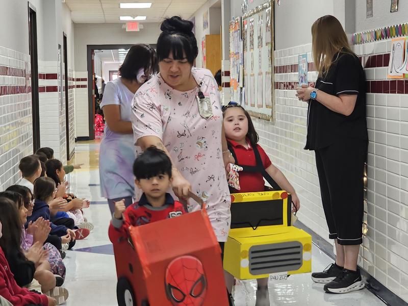 PK student with the Teacher Assistants guide 2 PK students on in a Spiderman car and the other in a school bus