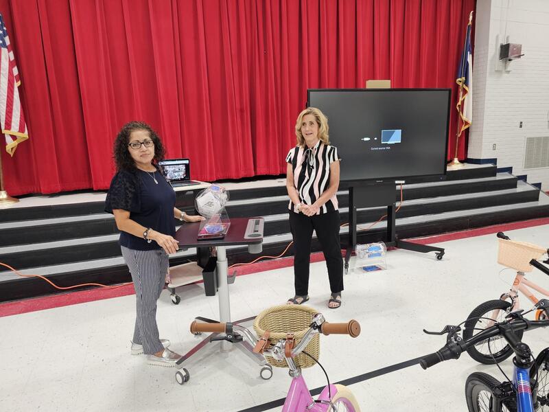 Mrs. Camack (Counselor) and Ms. Banegas (Principal) stand by the ticket dispenser. They are getting ready to take out the first winner's name. They stand in the gym in front of the stairs to the stage with the red curtain.