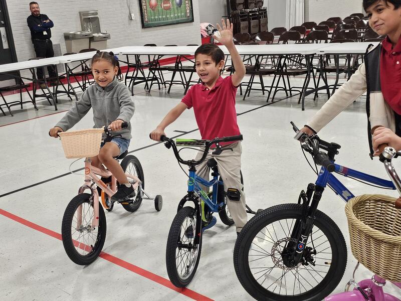 Kinder and 1st grade students smile and wave to the camera while sitting on their bikes