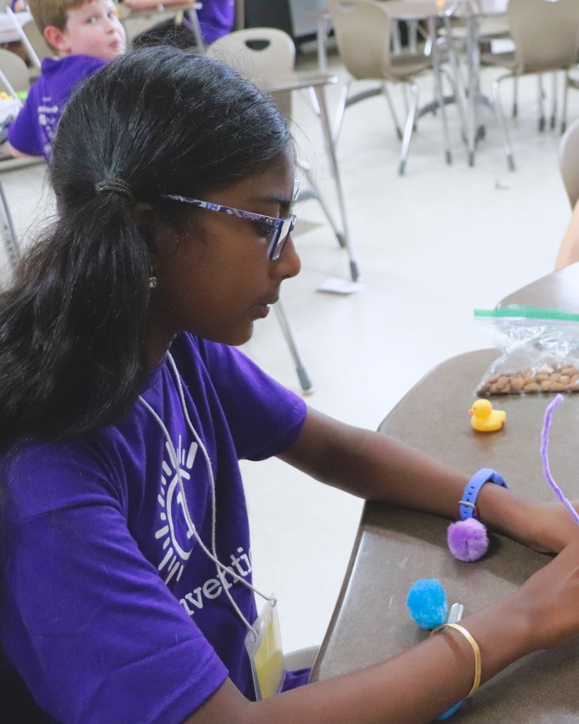 A student wearing glasses and a purple "Camp Invention" t-shirt sits at a desk, focused on a hands-on activity involving colorful craft materials. A plastic bag with beans and a small rubber duck are also on the desk. Other students are visible in the background, seated at desks in a classroom setting.