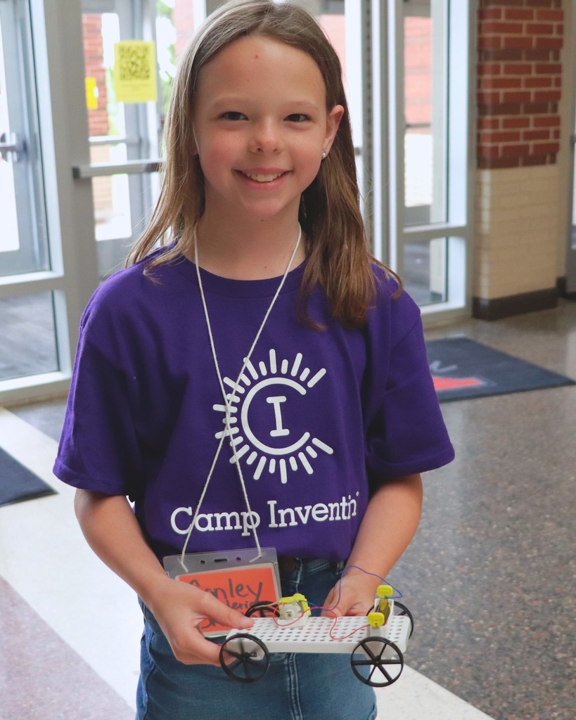 girl in purple shirt with a red name tag smiling and holding a car she made