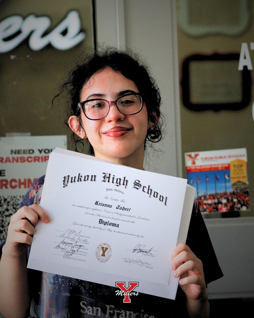 girl with a brown pony tail and black shirt holding up her white diploma and smiling