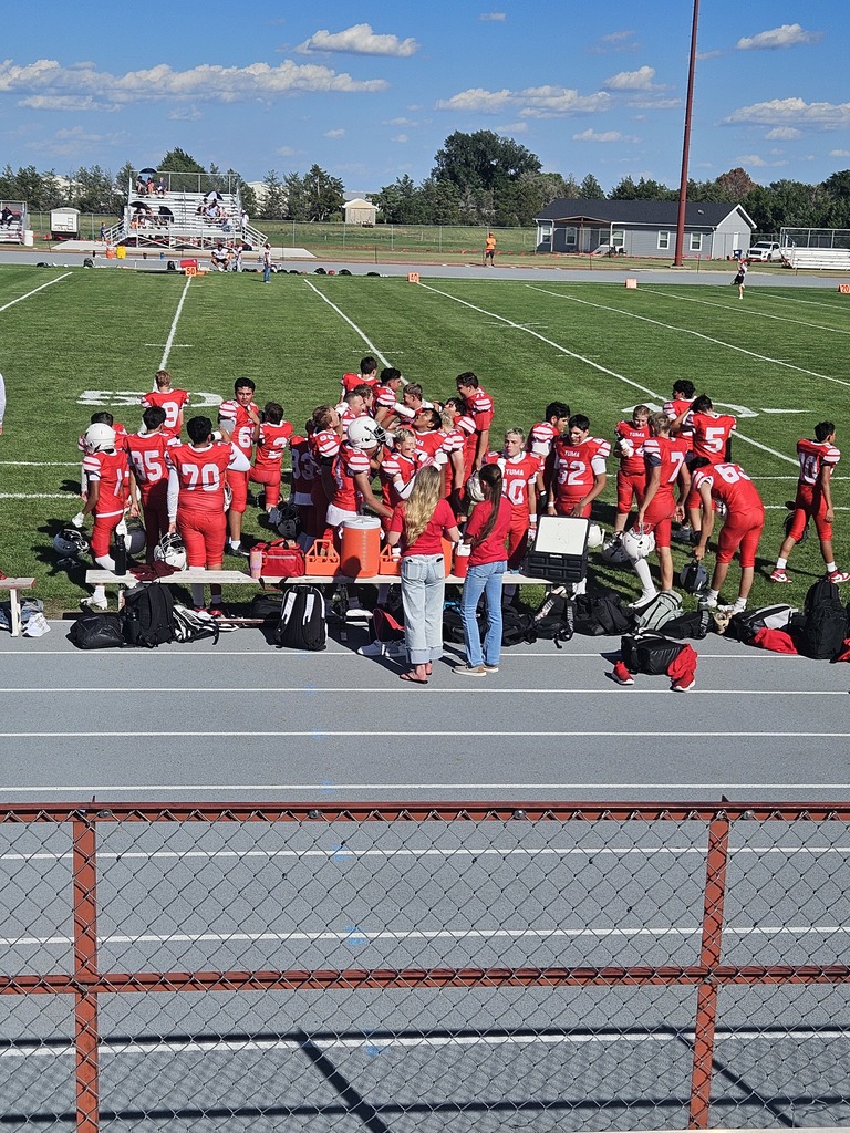Athletes in red uniforms and helmets stand around a table on a football field, with a fence in the foreground.
