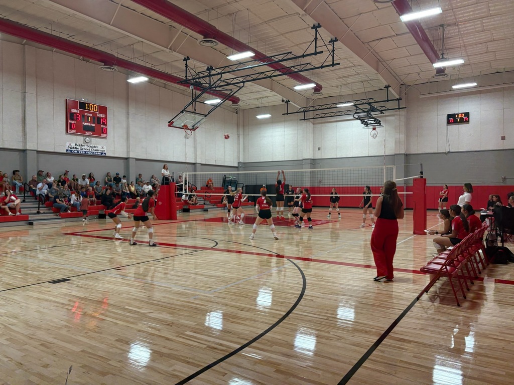An indoor gym with a wooden floor, red benches, and multiple people. A volleyball game is being played.