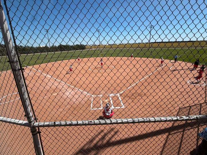 A baseball field with players in red and white uniforms, viewed from behind a metal chain-link fence.