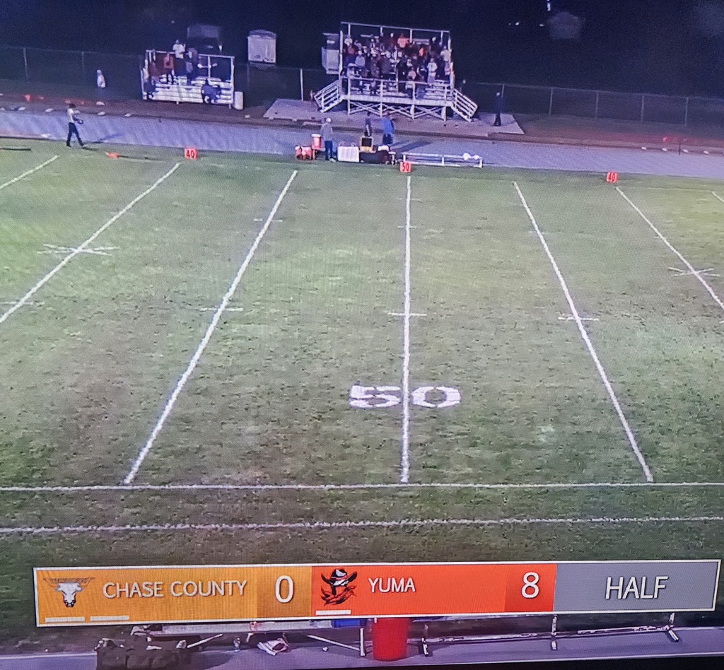 A football field with white lines, scoreboard showing Chase County 0, Yuma 8, and half. People in stands.