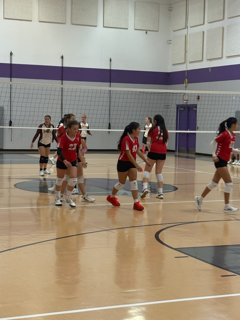 A group of players wearing red uniforms and shorts, playing volleyball in a gym with a wooden floor.
